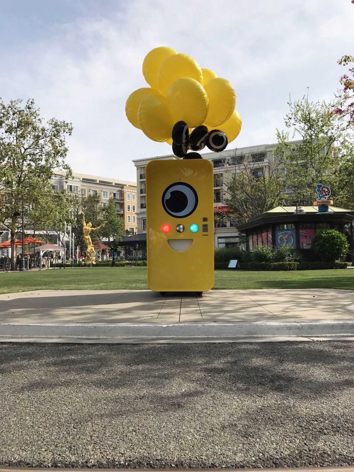 Yellow Snapbot vending machine with balloons on a lawn at an outdoor shopping center, pretzel kiosk and gold statue behind